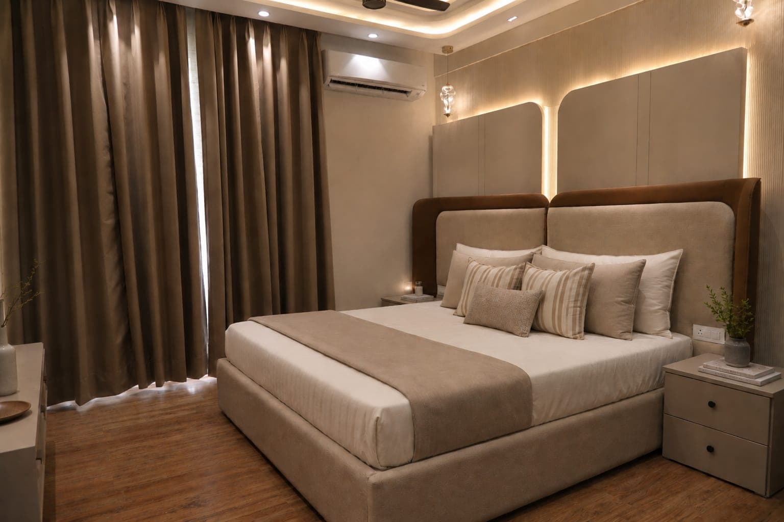 Bedroom with a cream upholstered bed framed by a brown wingback headboard, floor-length taupe curtains and warm cove lighting above a wood floor.
