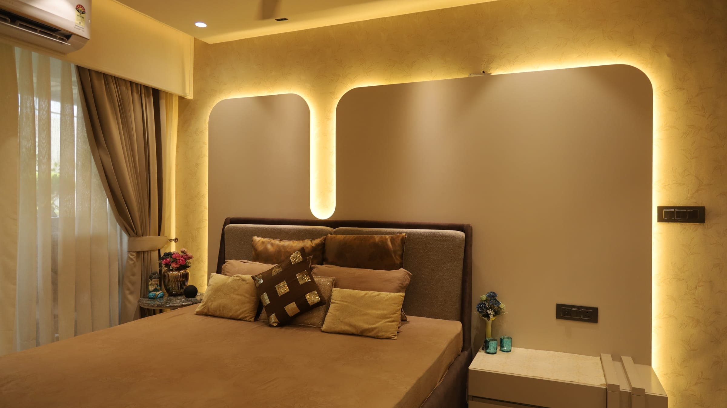 Bedroom with an upholstered bed, layered cushions and a curved headboard wall framed by warm concealed lighting, beside sheer and taupe curtains.