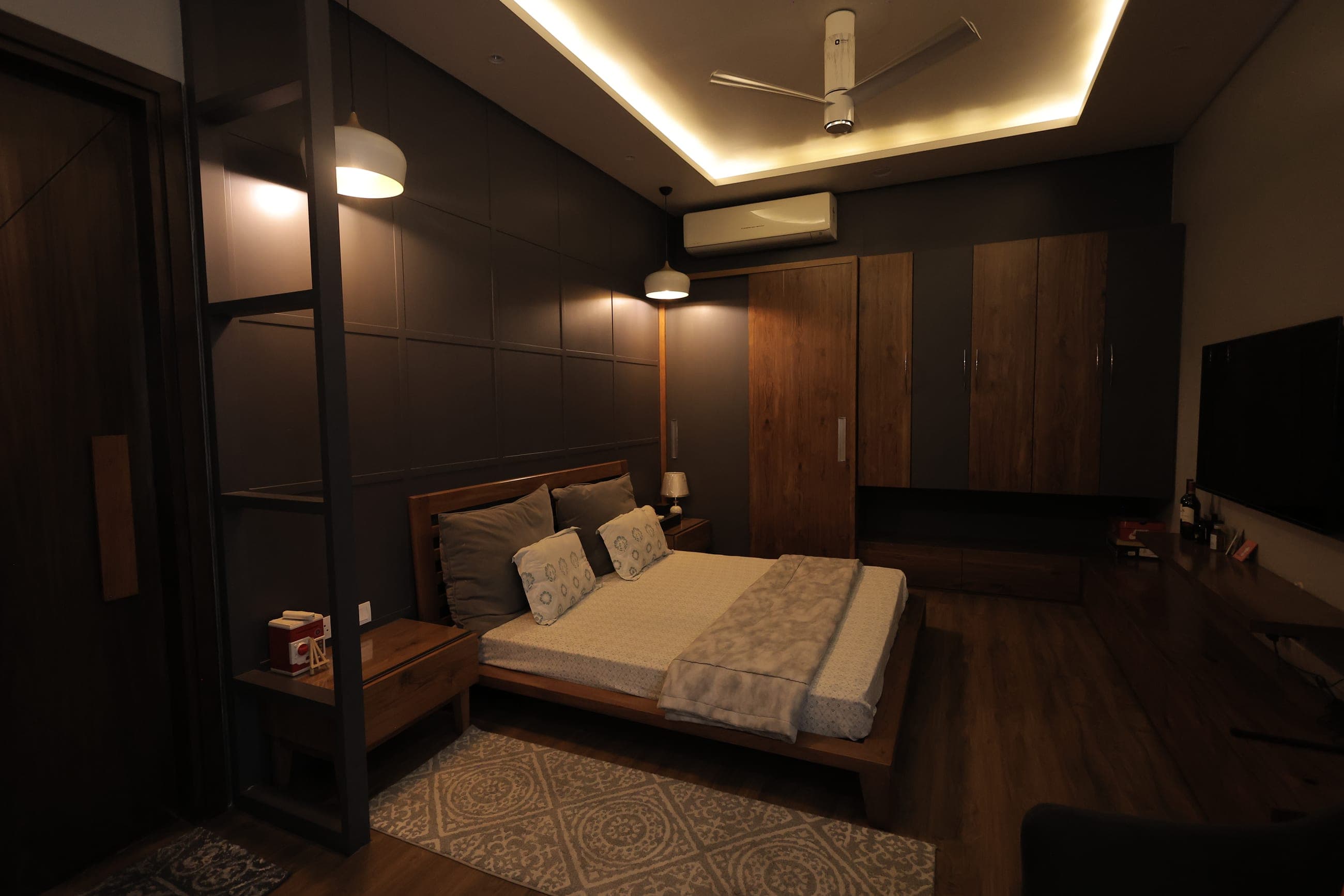 Bedroom at night with a wooden platform bed, dark panelled feature wall, warm cove lighting and a fitted timber wardrobe.
