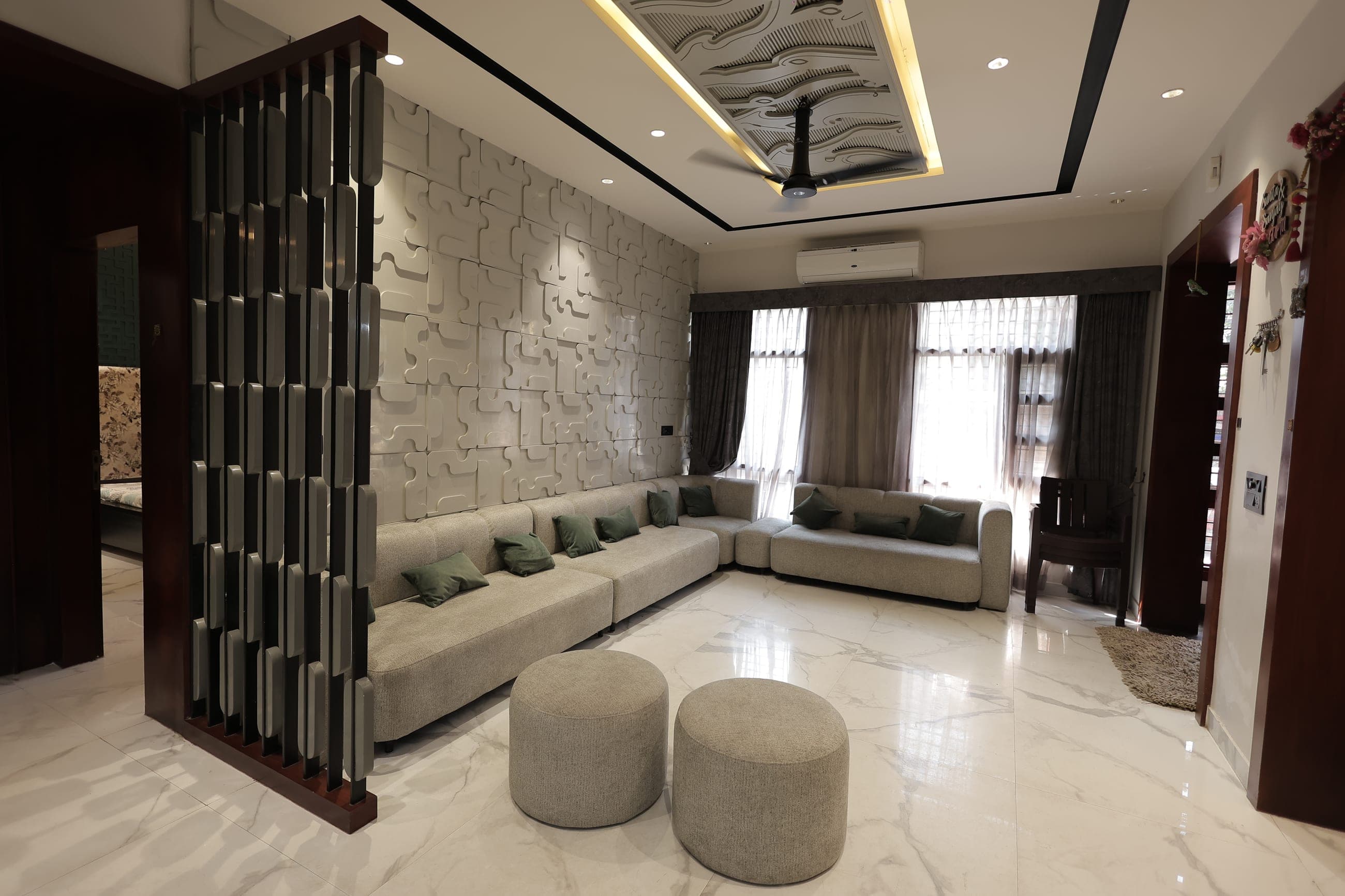 Living room with an L-shaped grey sofa and green cushions against a sculpted three-dimensional white feature wall, a marble floor, two round fabric ottomans and a dark slatted wood partition.
