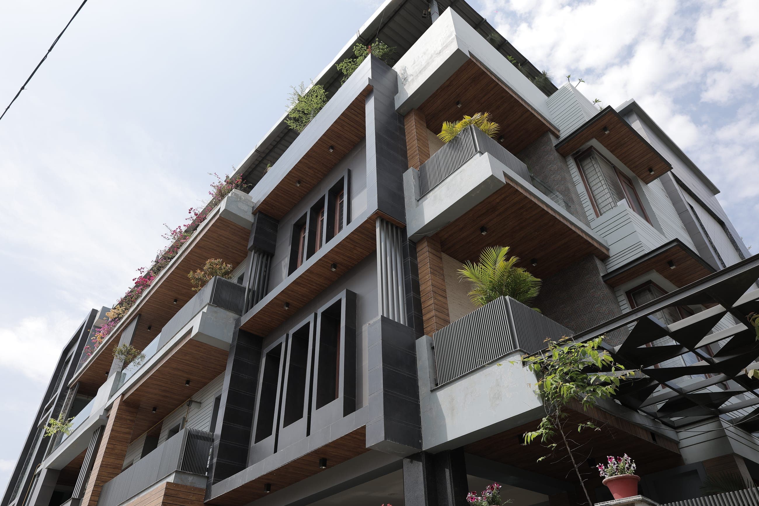 Low-angle view of a modern multi-storey house corner, with dark grey stone cladding, warm timber panels, slatted metal balcony screens and rooftop greenery against a bright blue sky.