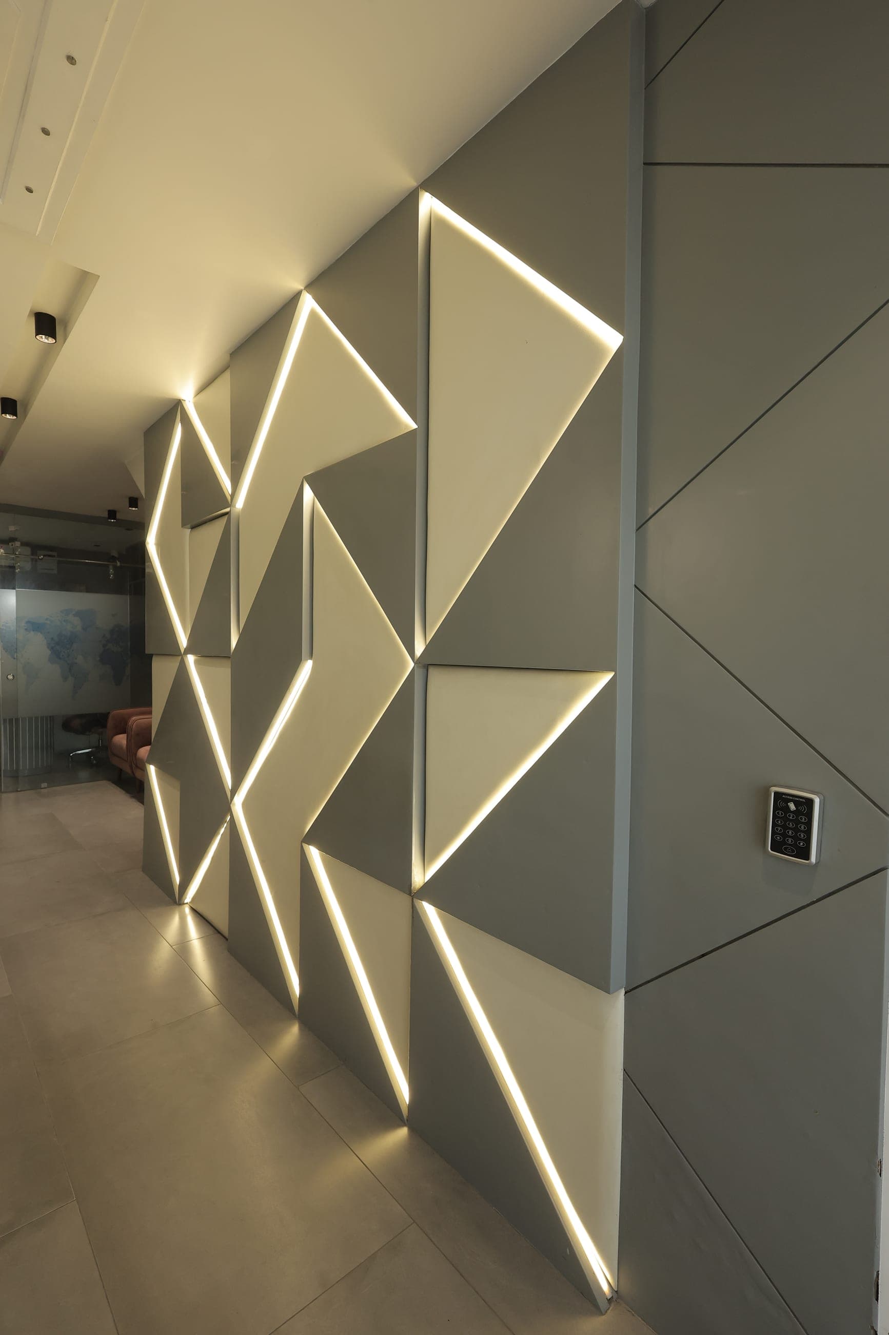Backlit feature wall of angled triangular panels edged with warm LED strip lighting, beside pale stone tiling, a keypad access panel and a glass partition.