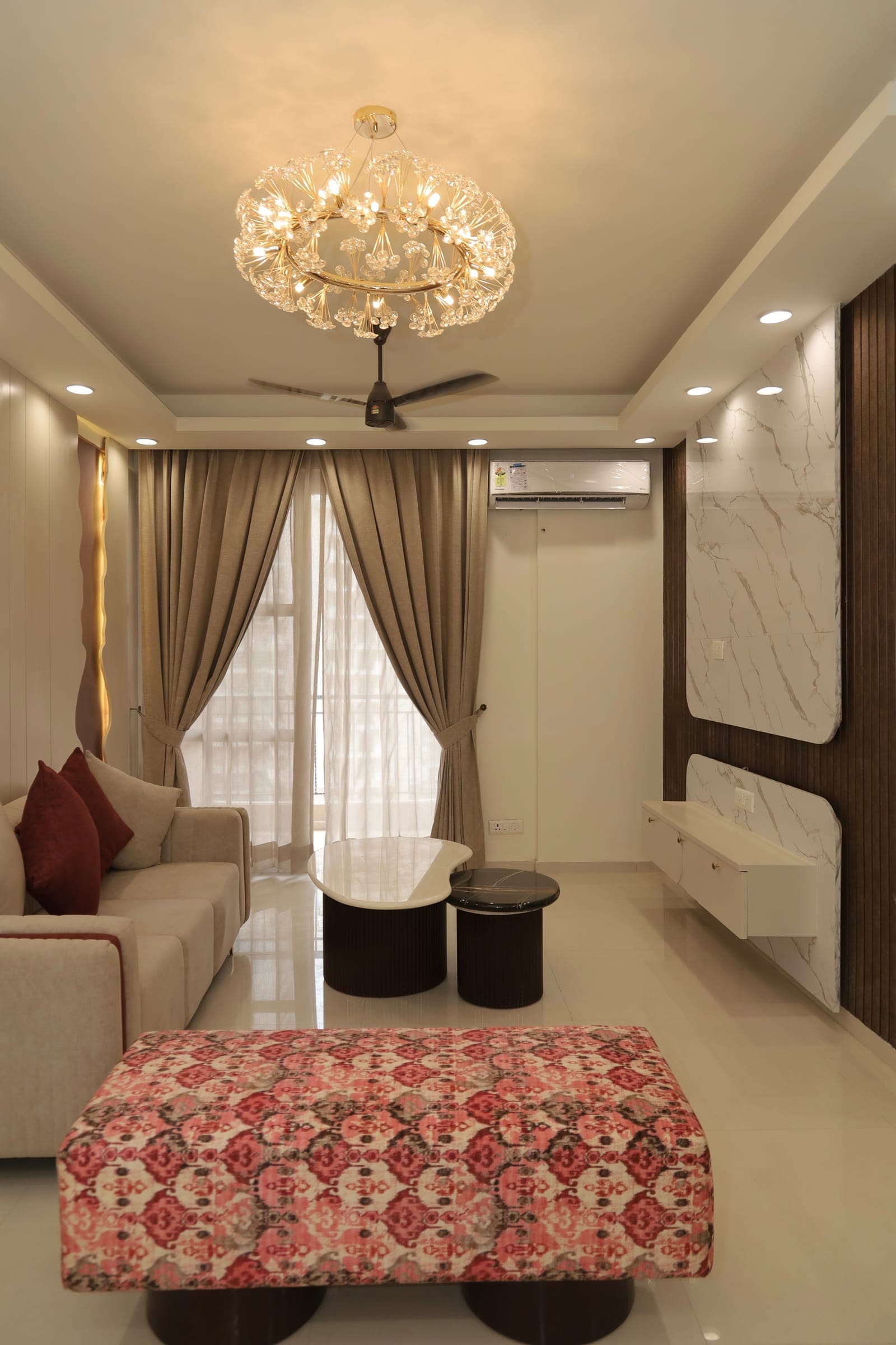 Living room with a beige sofa, a red-and-cream patterned ottoman, a floral crystal chandelier and a marble-panelled feature wall.