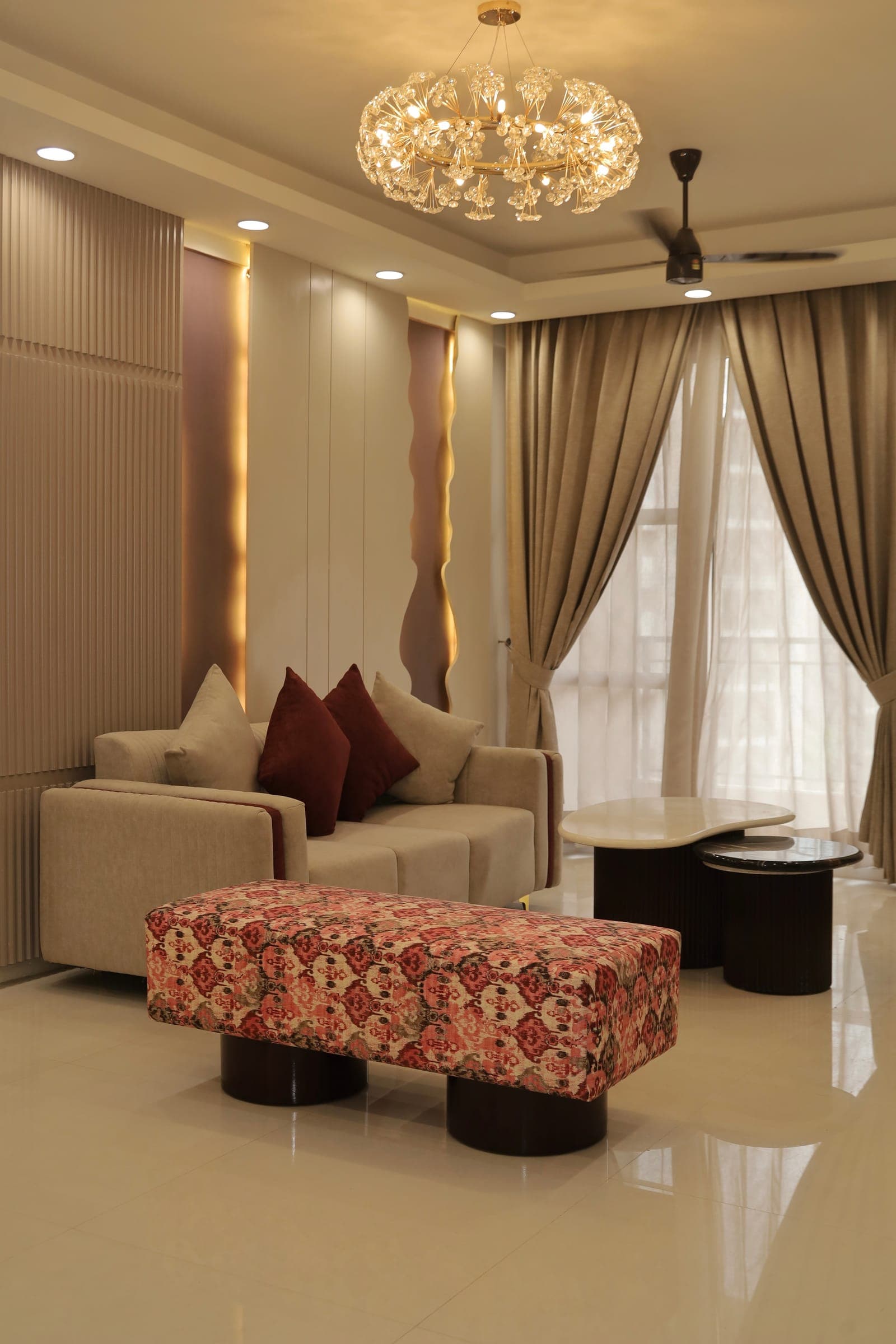 Living room seating with a beige sofa, maroon and grey cushions, a patterned bench and sheer curtains beside a tall window.