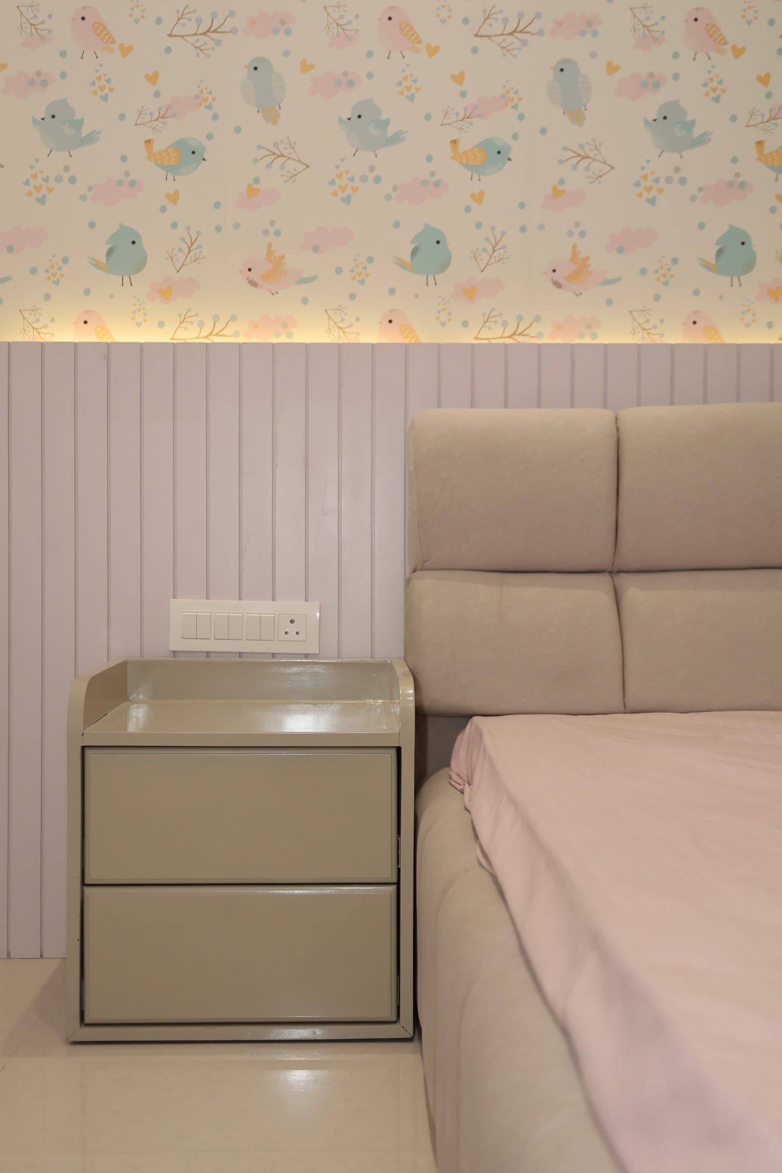 Children's bedroom corner with pastel bird wallpaper, fluted panelling, a sage bedside cabinet and a cushioned headboard.