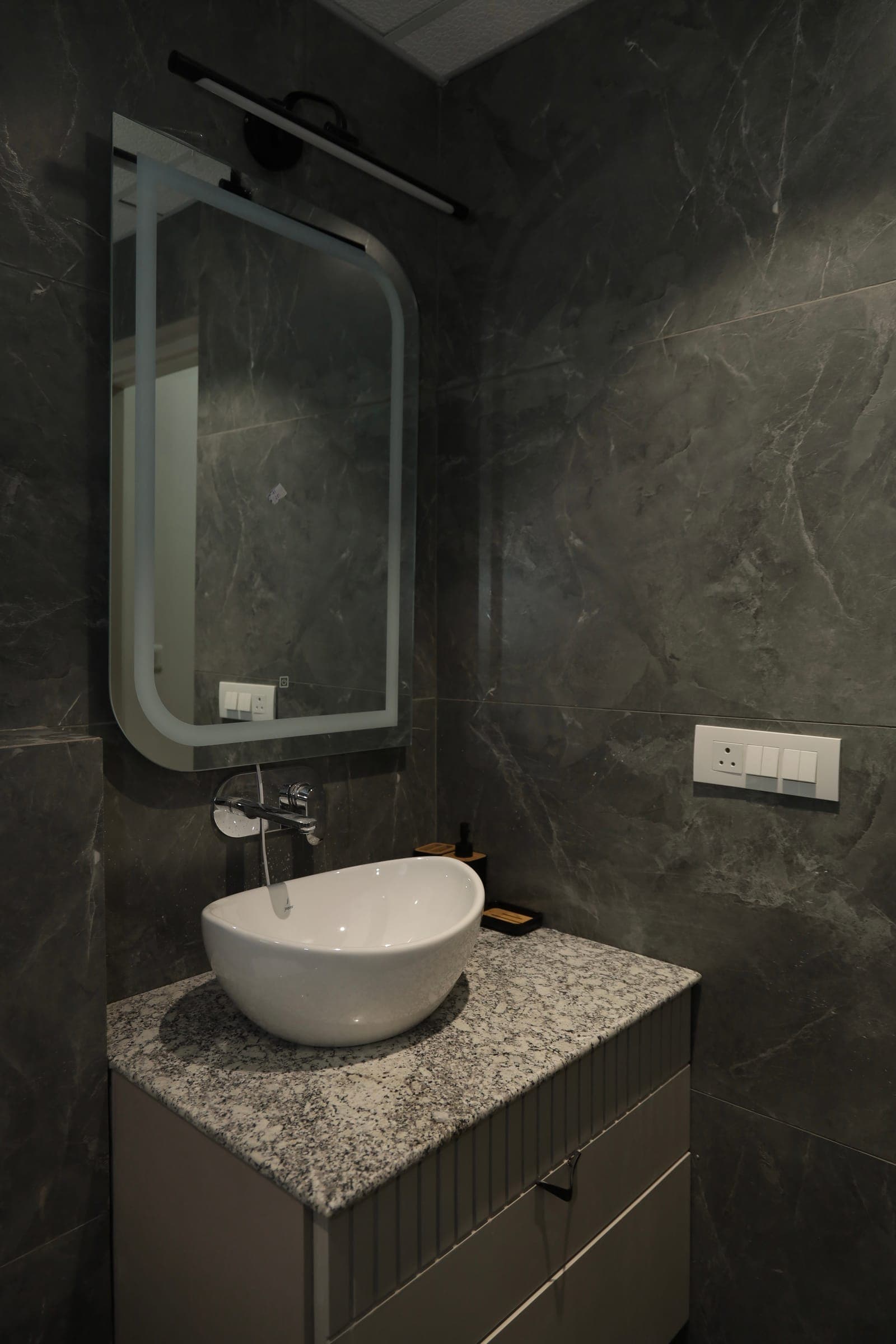 Washroom with dark grey marble-look walls, a white vessel basin on a speckled stone top and a backlit mirror.