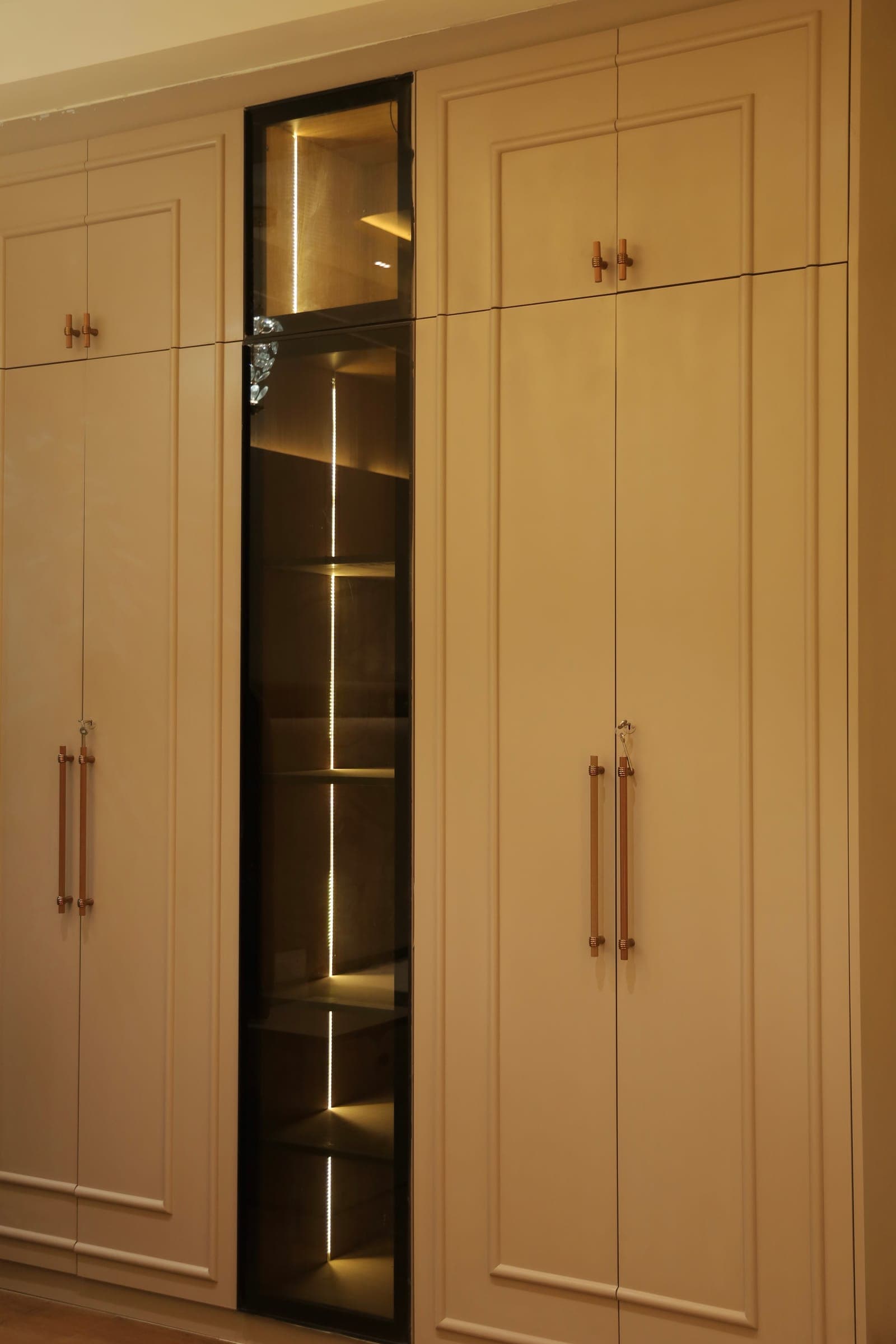 Built-in wardrobe with cream panelled doors, slim copper handles and a lit glass display column.