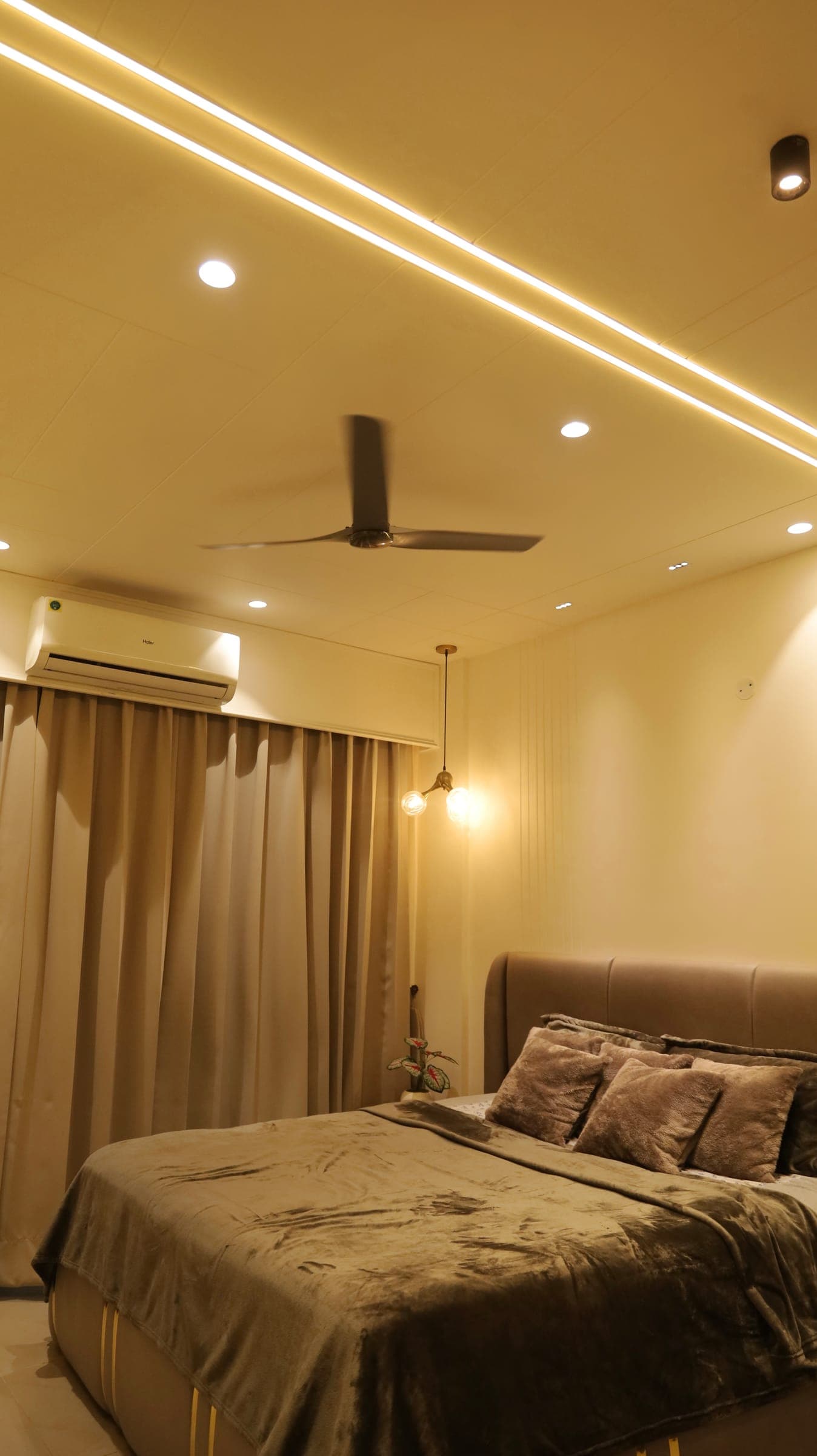 Bedroom with an upholstered bed and grey throw, a two-globe pendant light, a ceiling fan, recessed lights and floor-length curtains beside a wall air-conditioner.