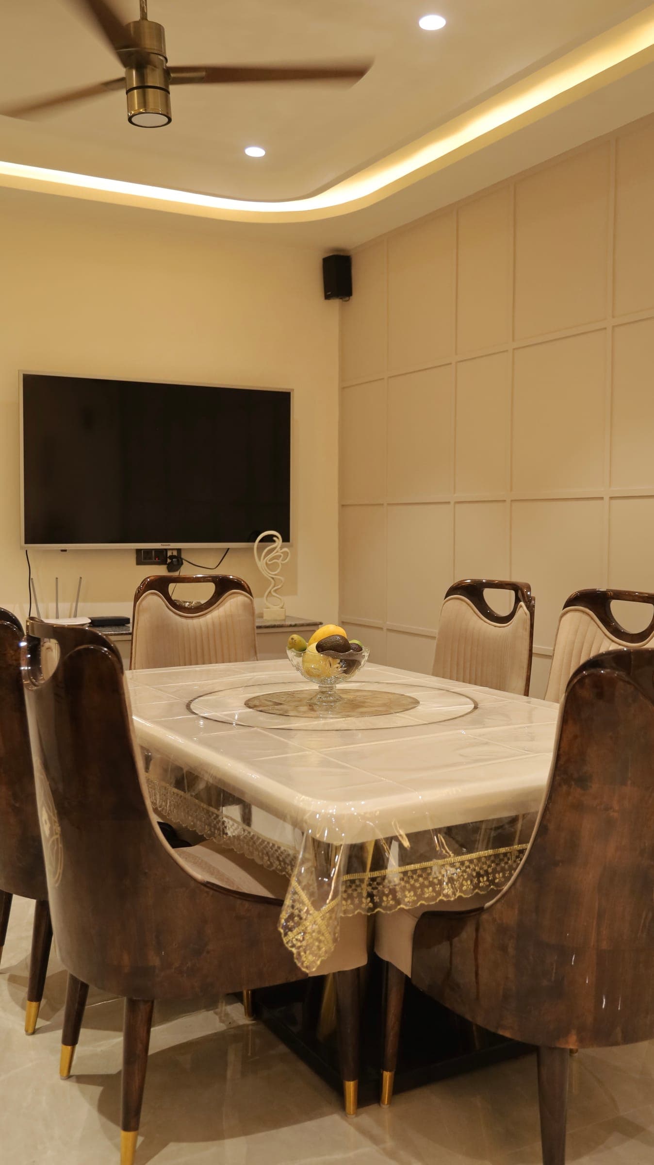 Dining area with a marble-top table and six upholstered chairs, a fruit bowl at the centre, a wall-mounted TV and a panelled cream wall.