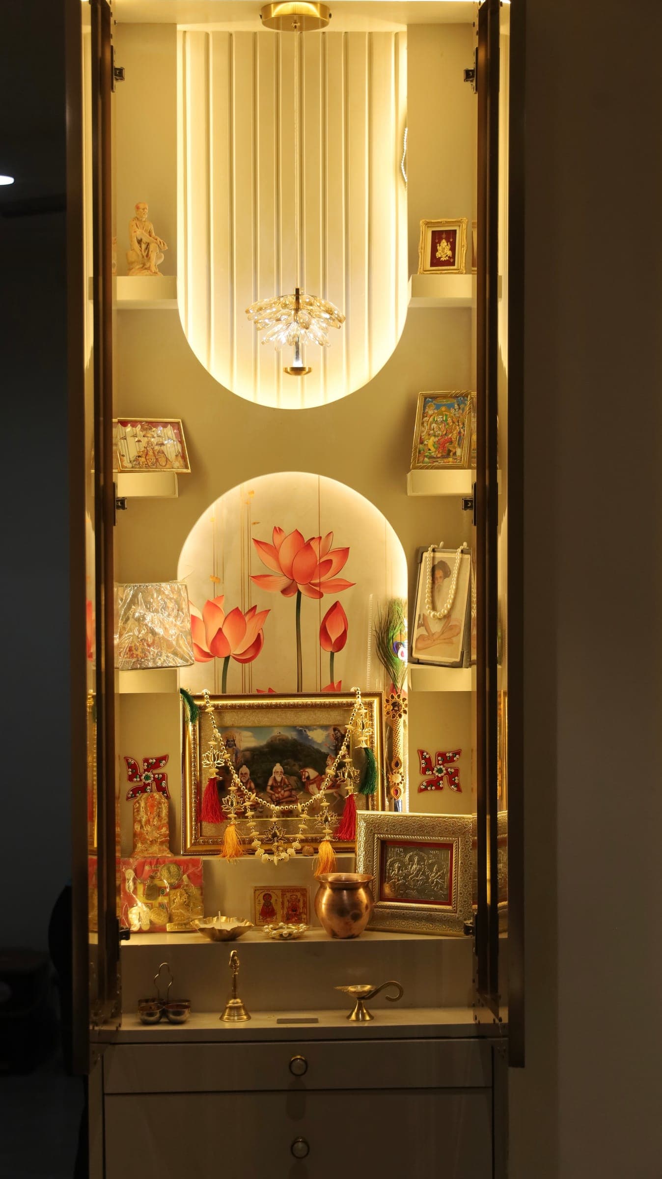 Prayer niche with backlit fluted panelling, a lotus mural, framed devotional pictures and brass lamps on a white counter.