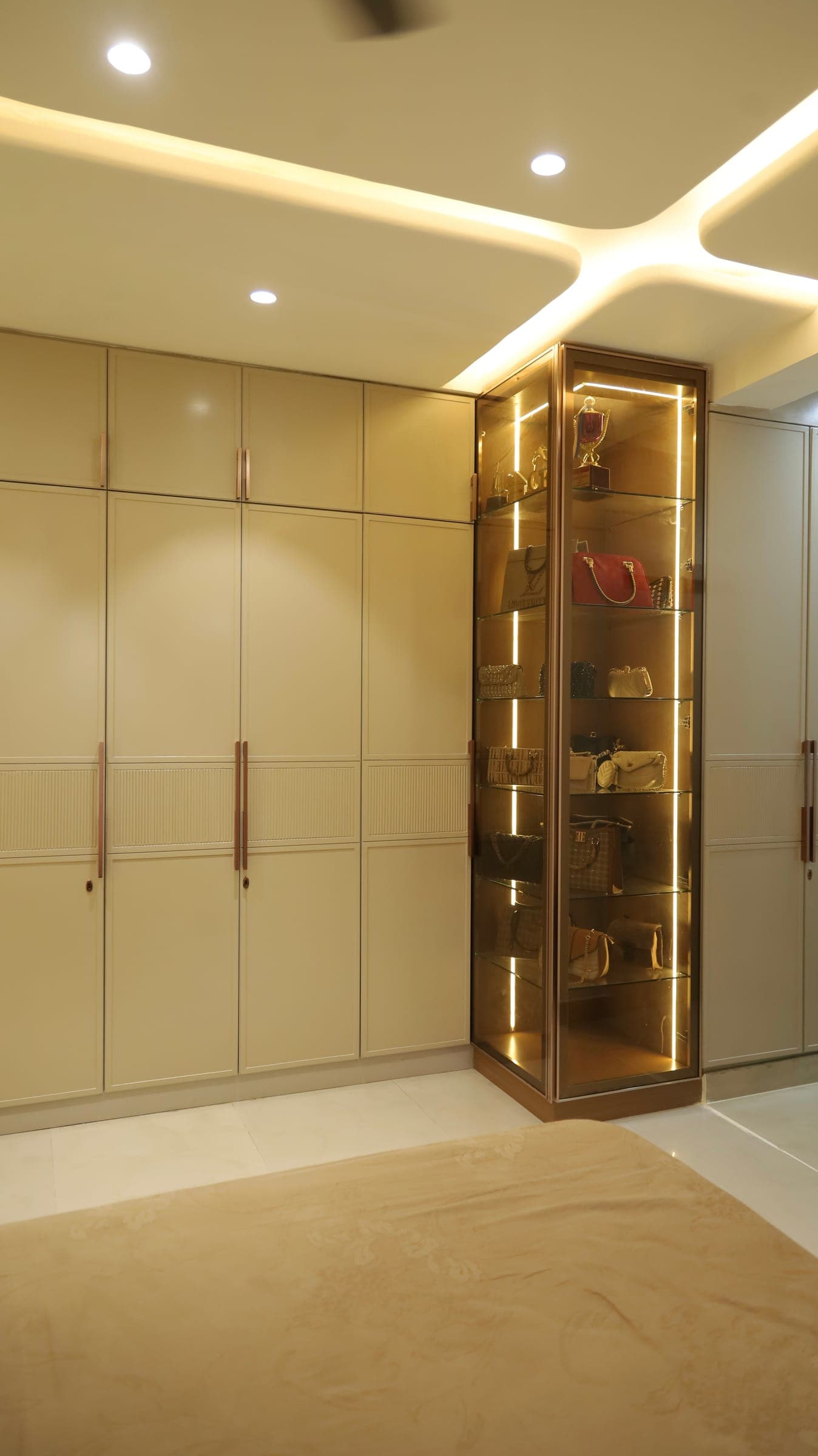 Dressing area with floor-to-ceiling cream wardrobes and a corner glass display cabinet, lit from within, holding handbags and decorative pieces.