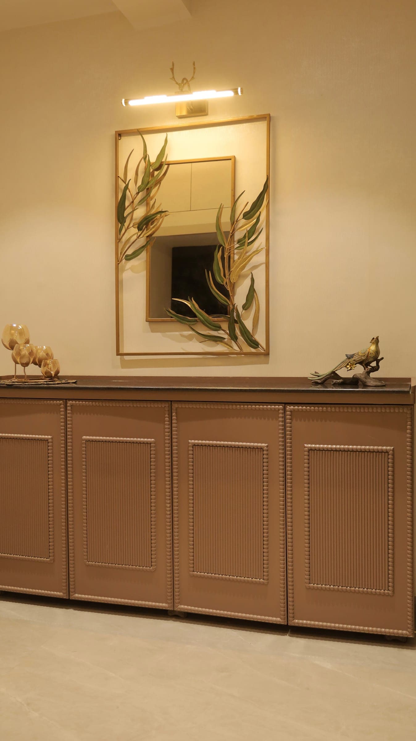 Console with a dark stone top and beaded panelled doors, styled with glass goblets and a bird figurine below a leaf-framed mirror and a brass picture light.