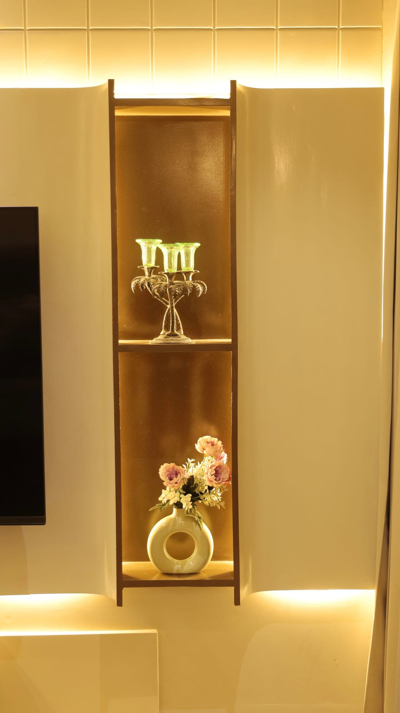Backlit wall niche holding a silver palm-shaped candelabra with green glass cups and a round white vase of flowers, beside a wall-mounted TV.