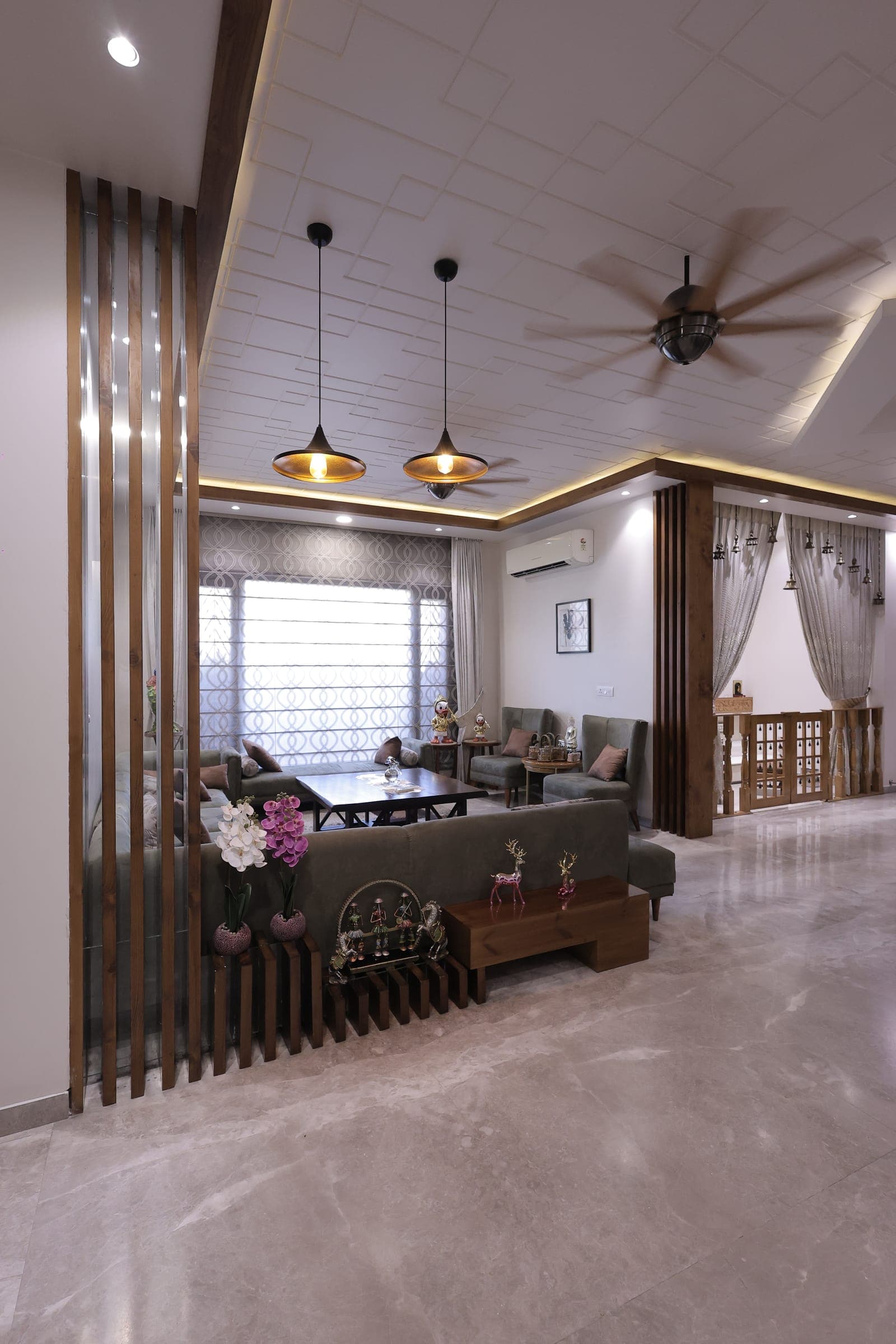 Living room with a grey velvet sofa, a wooden console with flowers and decor, a sheer-curtained window and twin gold pendant lights below a patterned false ceiling.