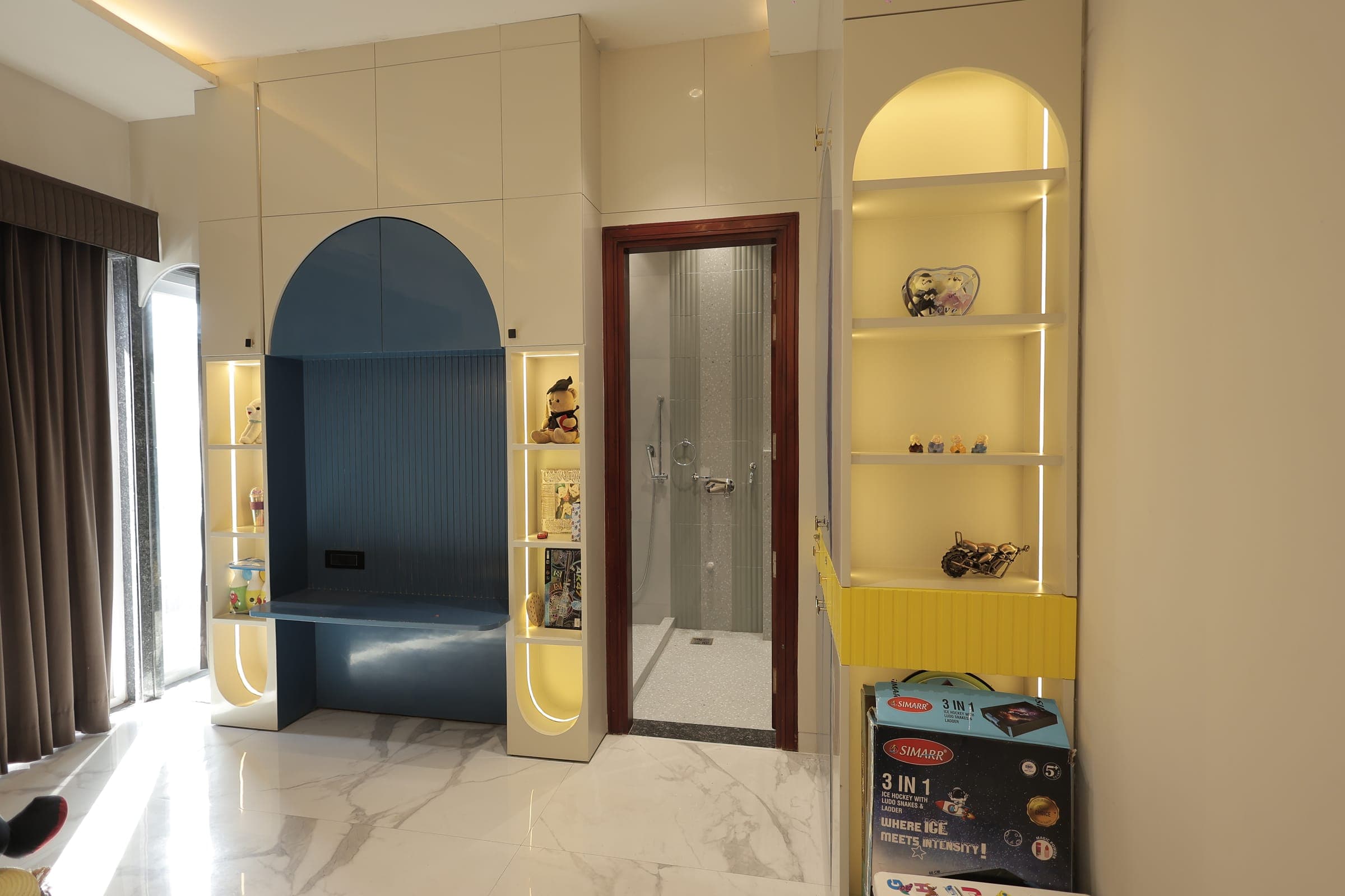 Child's room with an arched blue study nook, lit display niches holding soft toys and books, a glossy marble floor and an attached shower behind a wood-framed door.