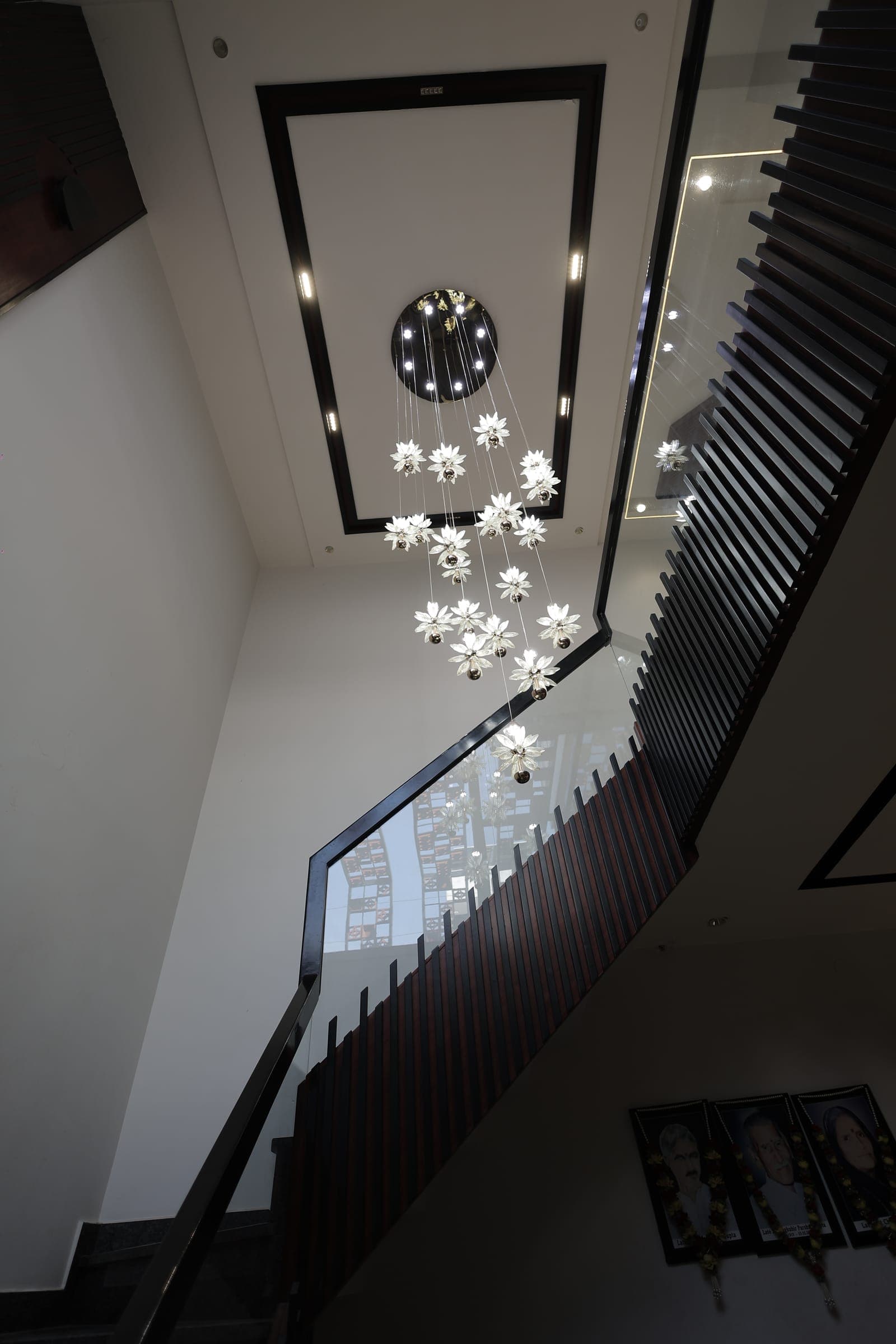 Stairwell with dark slatted wood railings and a tall cascading chandelier of glass flowers strung against a white double-height wall.