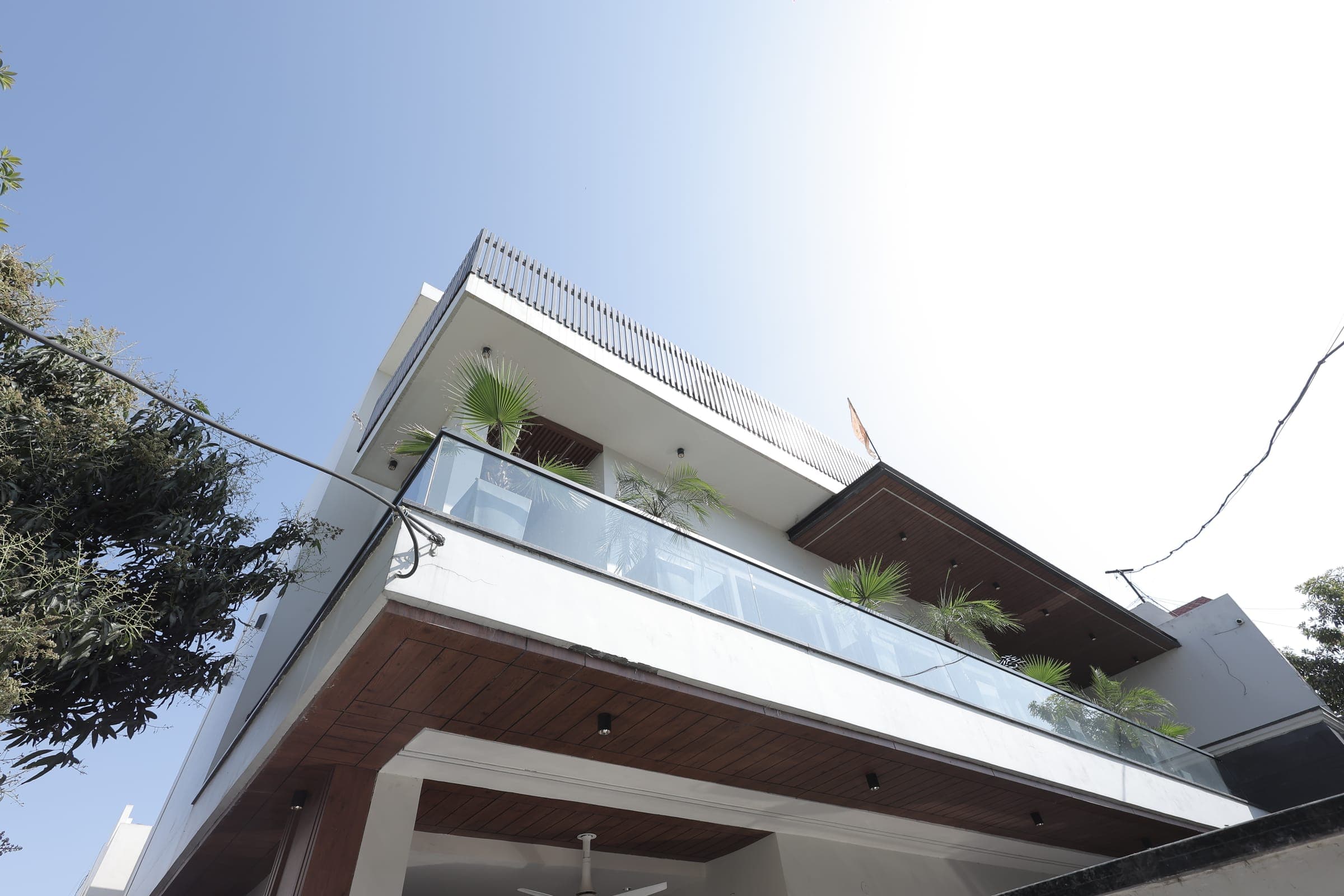 Low-angle view of the house exterior showing cantilevered balconies, a wood-slatted parapet, glass railings and potted palms against a bright sky.