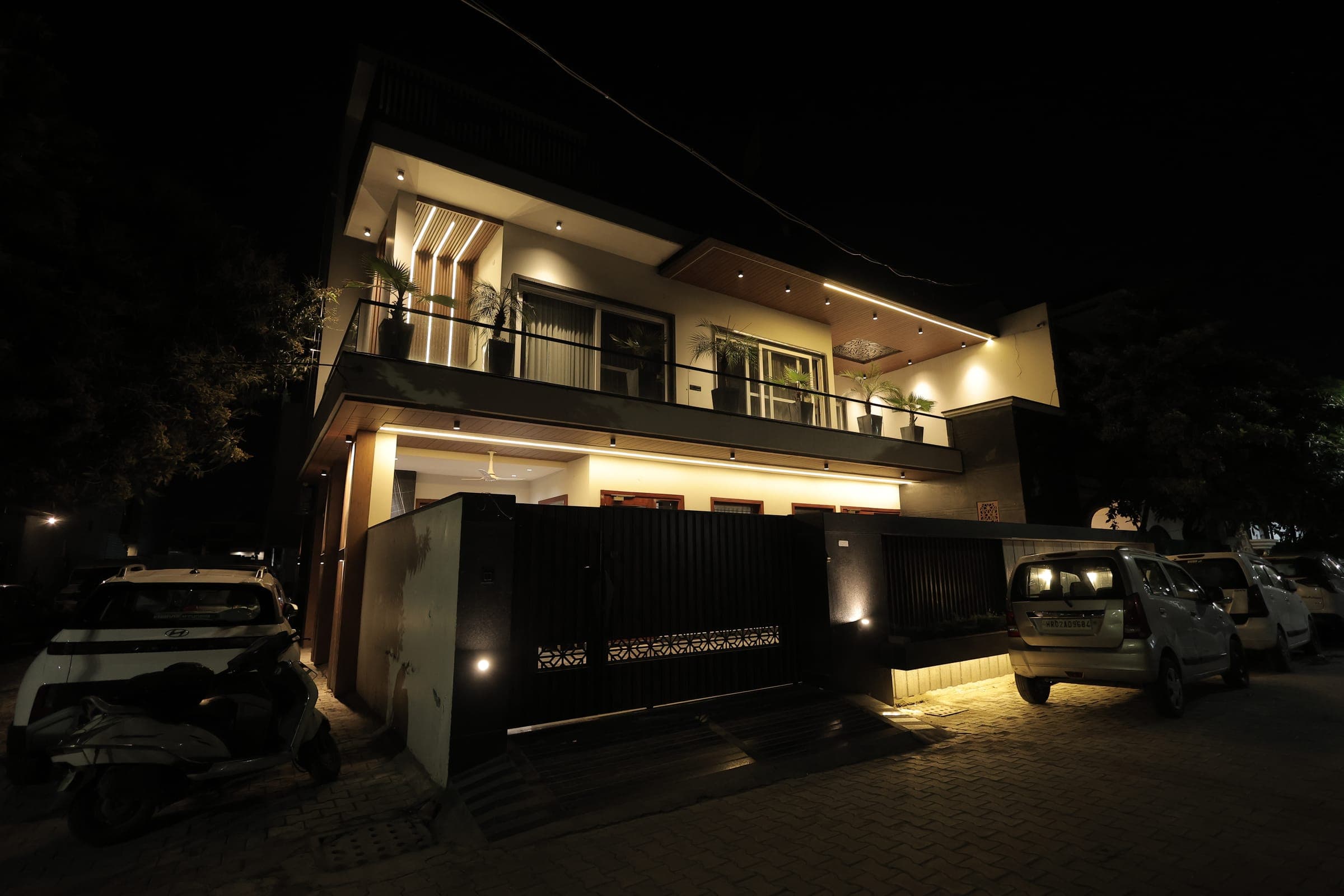 The house exterior at night with warm facade and soffit lighting, a wood-slatted entrance gate, balcony palms and cars parked outside.