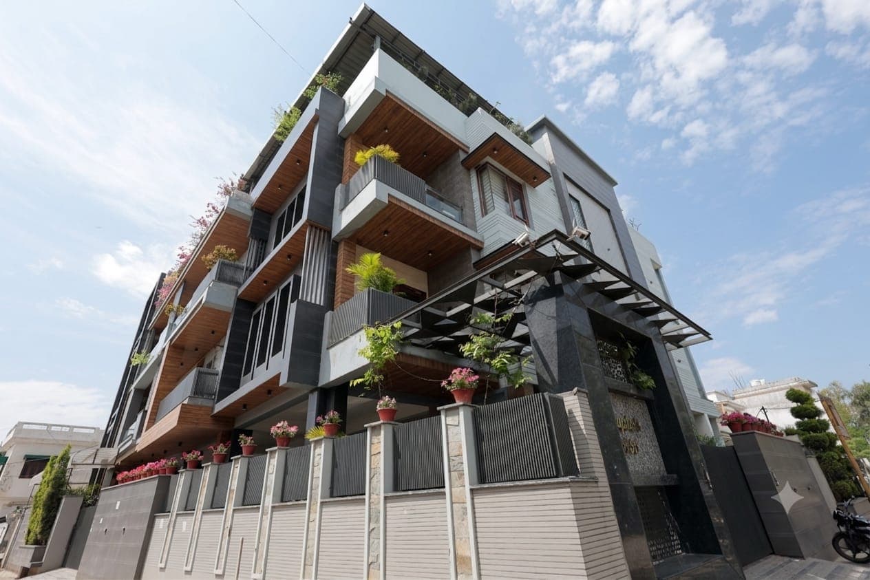Three-quarter street view of a contemporary three-storey home on a corner plot, combining grey stone, timber cladding and glass balconies, with a steel pergola and potted flowers along the boundary wall.