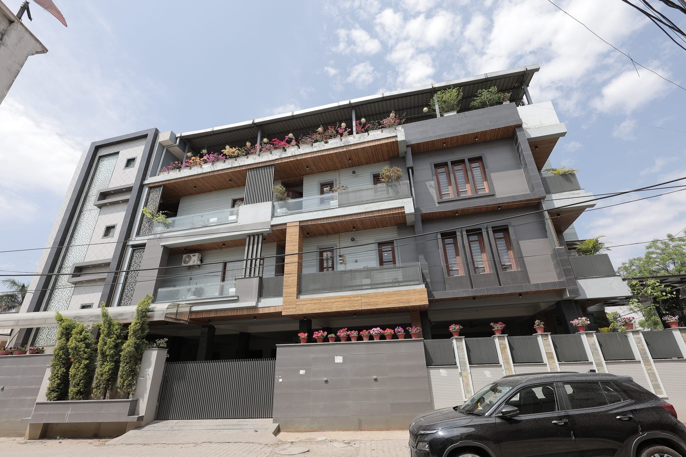 Full front elevation of a modern grey and timber multi-storey house, with glass-railed balconies, wooden windows, a flowering roof terrace and rows of potted plants above the compound wall.