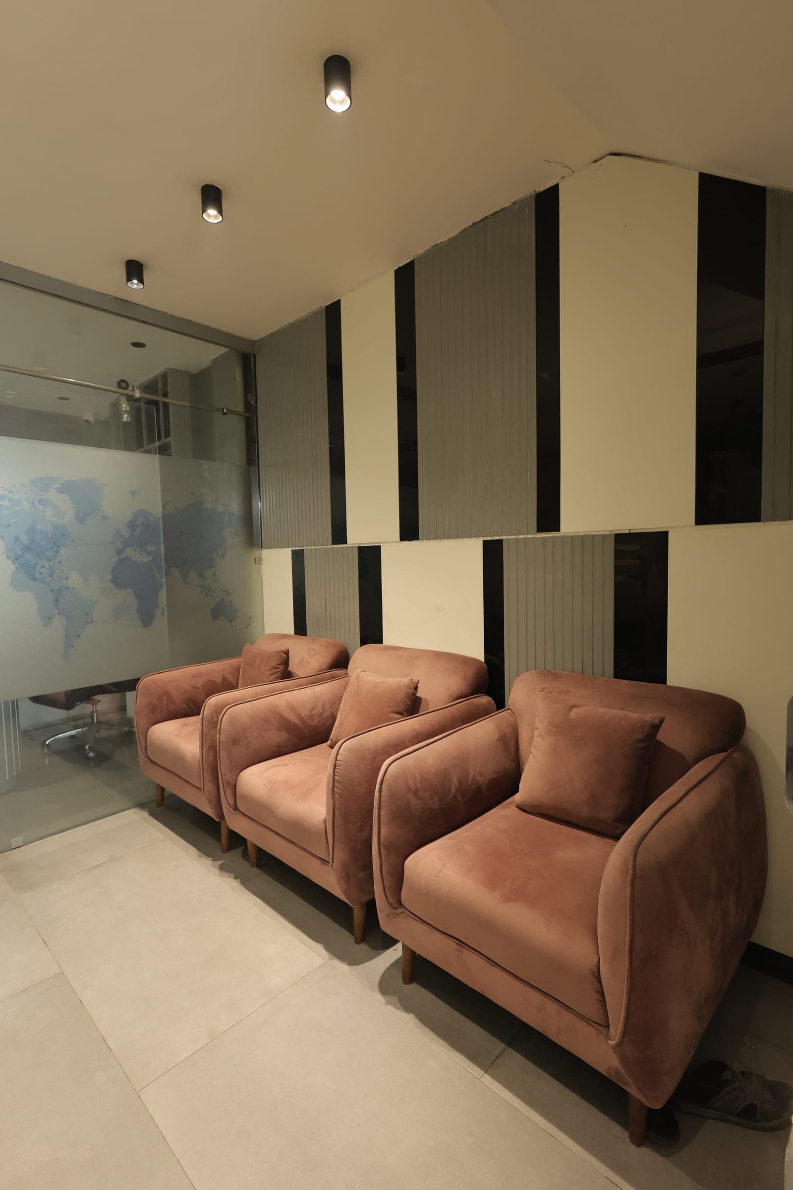 Row of dusty-rose velvet lounge chairs along a stone-tiled wall, with a black-and-white striped floor, cylindrical wall lights and a frosted glass partition behind.