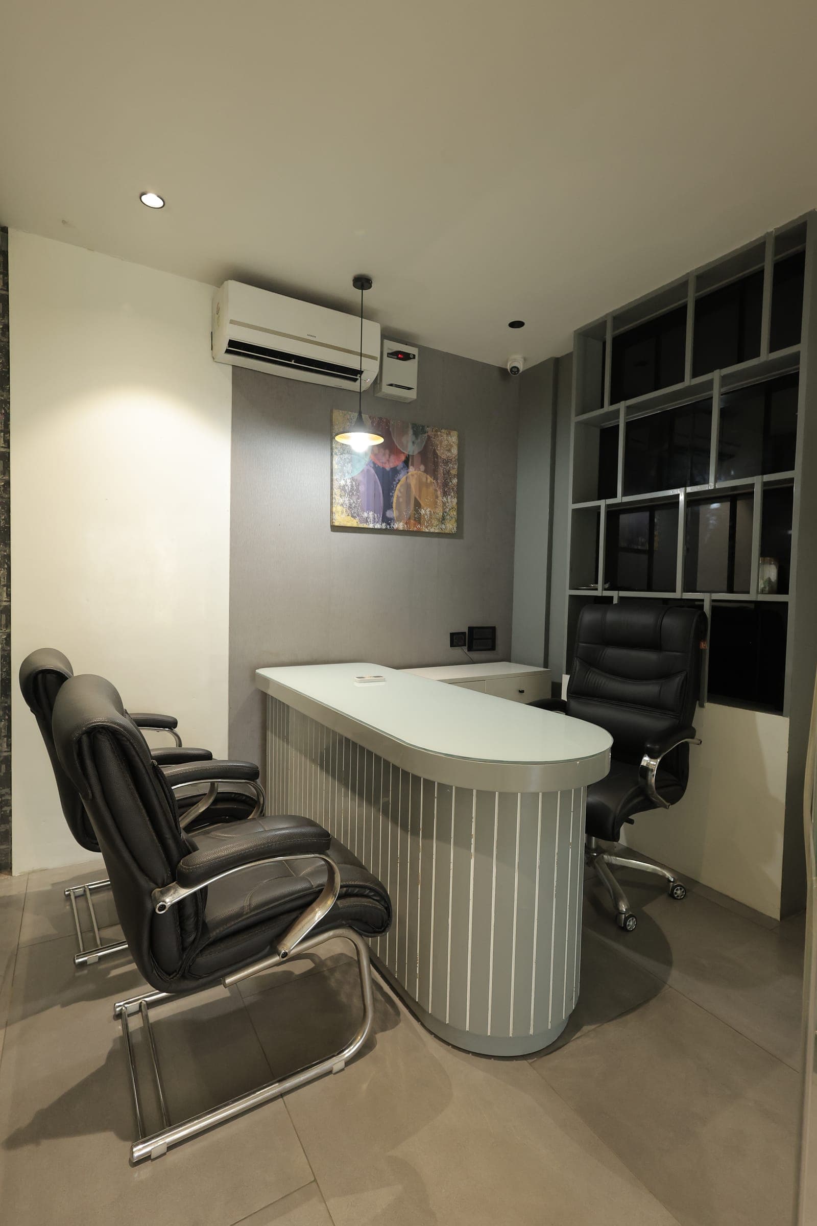 Office cabin with a curved slatted-front executive desk, black leather chairs, a wall-mounted air conditioner and a framed abstract print on a grey wall.